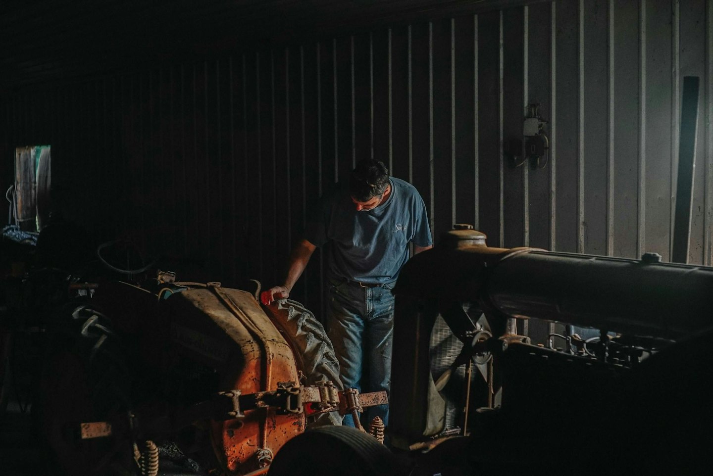 Man inspects an old rusty orange tractor in a barn.